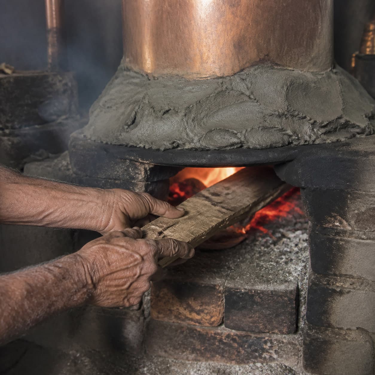An artisan feeding firewood into the brick furnace beneath a copper deg